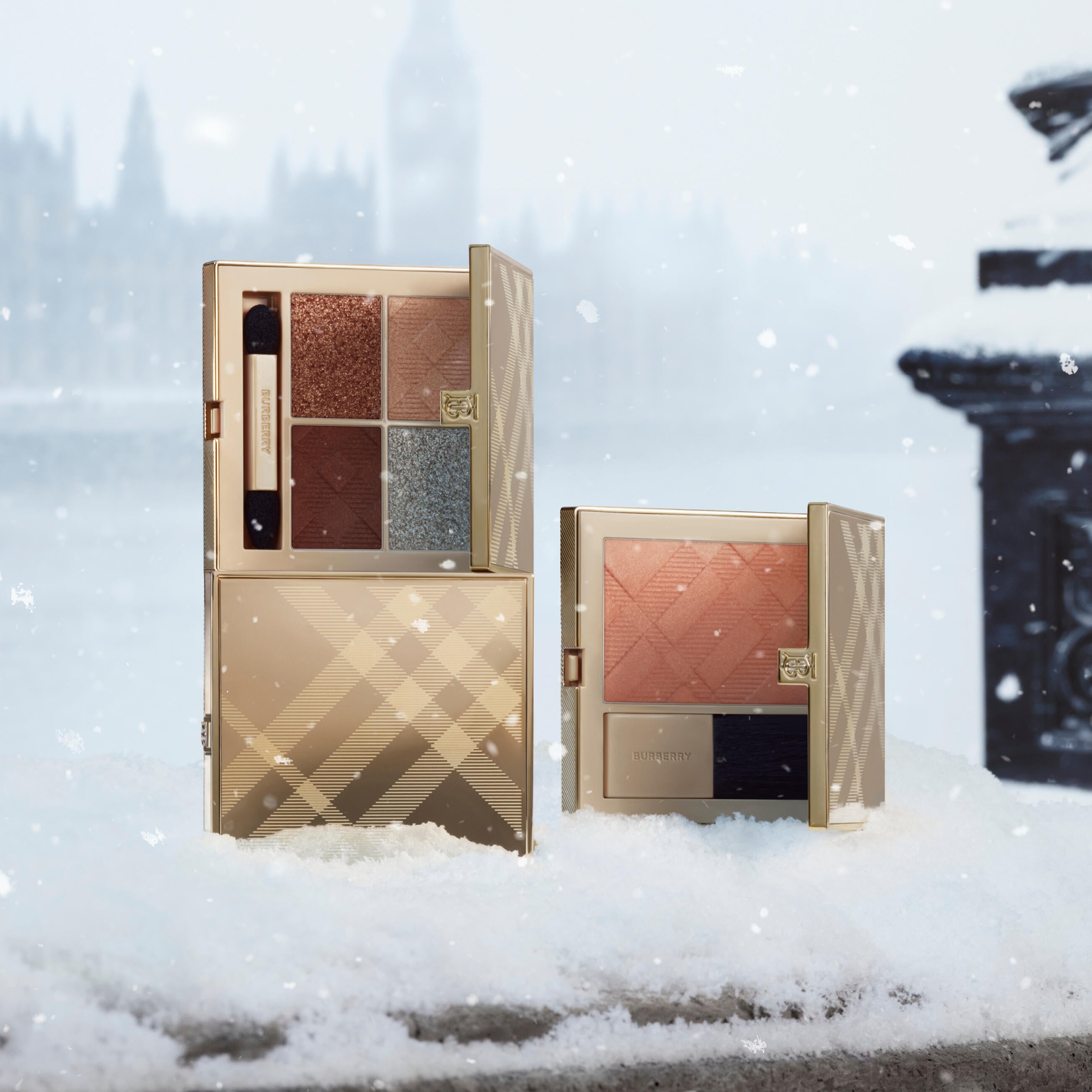 Gold Burberry check compacts standing in snow: an open four-colour eye palette, a closed case and an open peach-toned blush beside its brush.