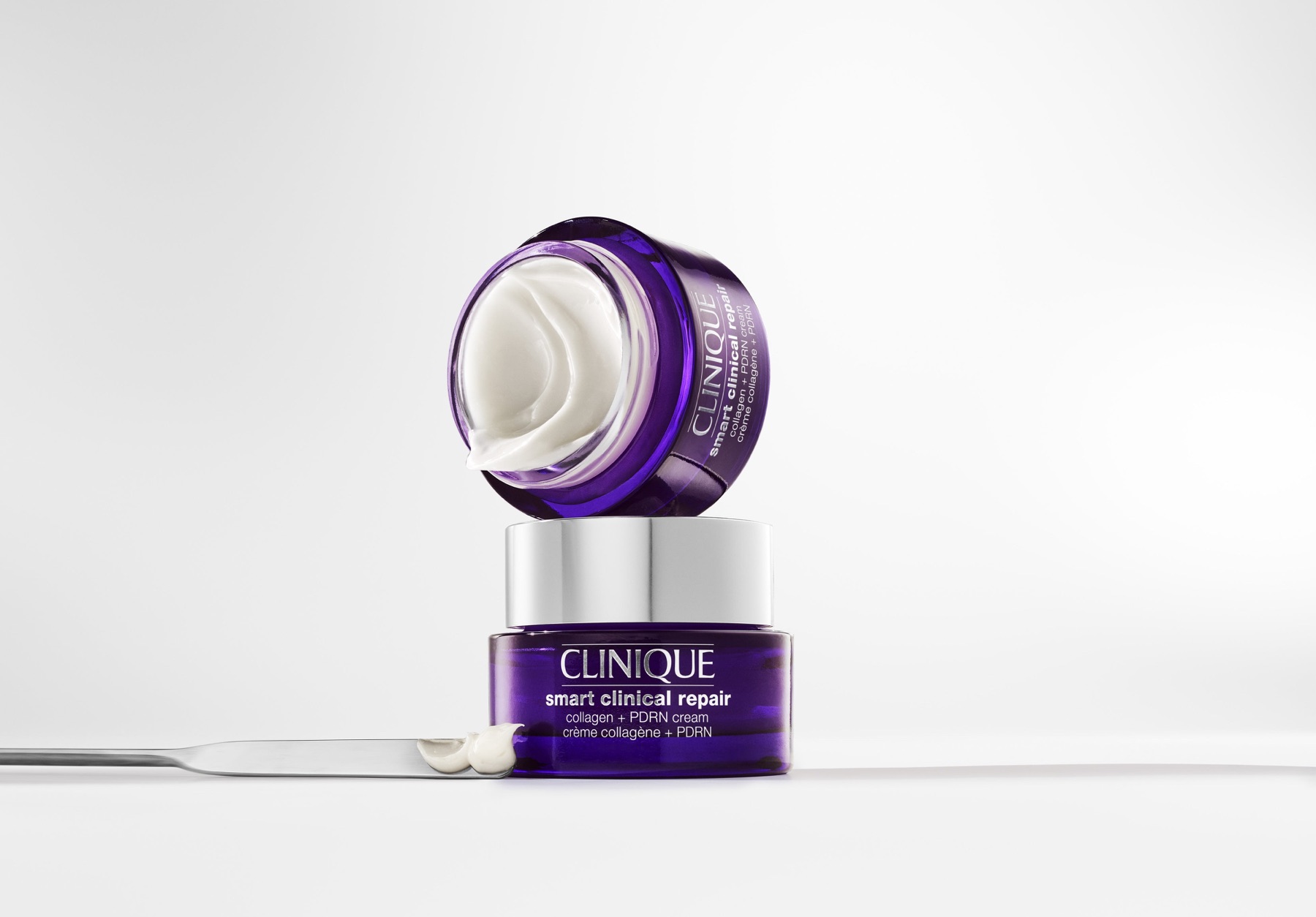 Two purple Clinique Collagen + PDRN Cream jars, one open and tilted above the other, beside a silver spatula on a pale background.