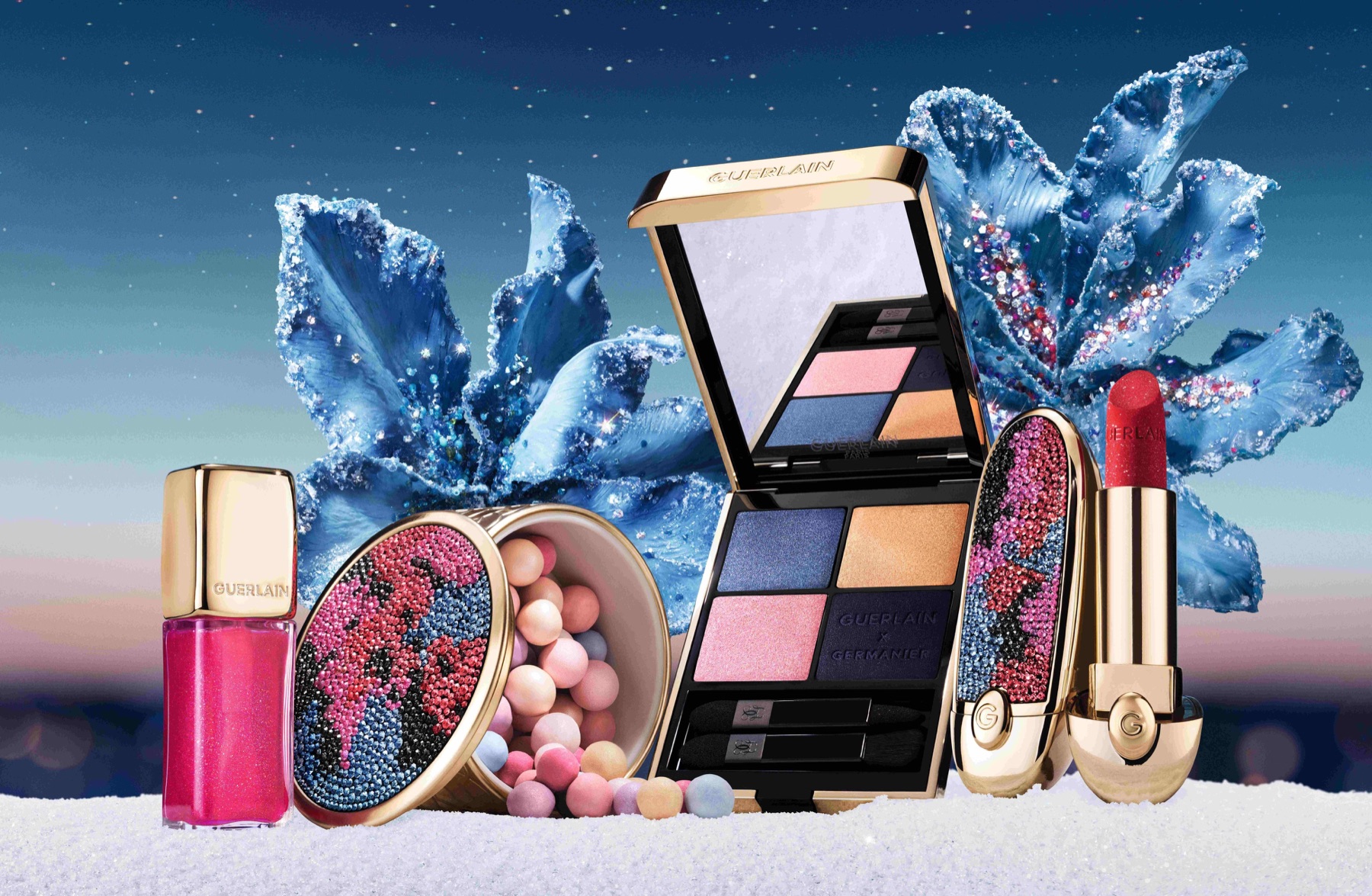 Open Guerlain eye palette and powder pearls beside a pink lip oil and red lipstick, with rhinestone-covered cases and blue embellished flowers.