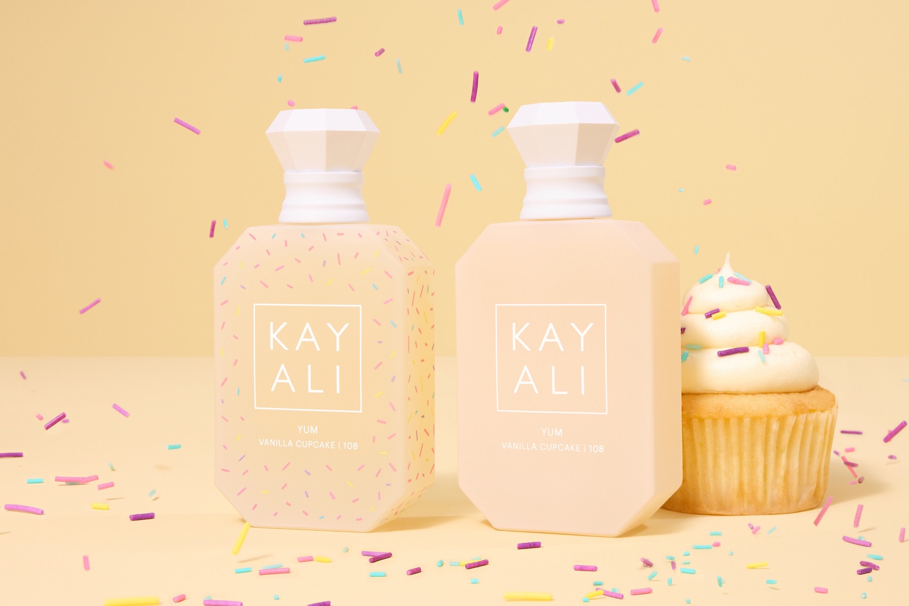 Two cream-coloured KAYALI Yum Vanilla Cupcake perfume bottles, one decorated with sprinkles, beside a frosted cupcake on a yellow background.