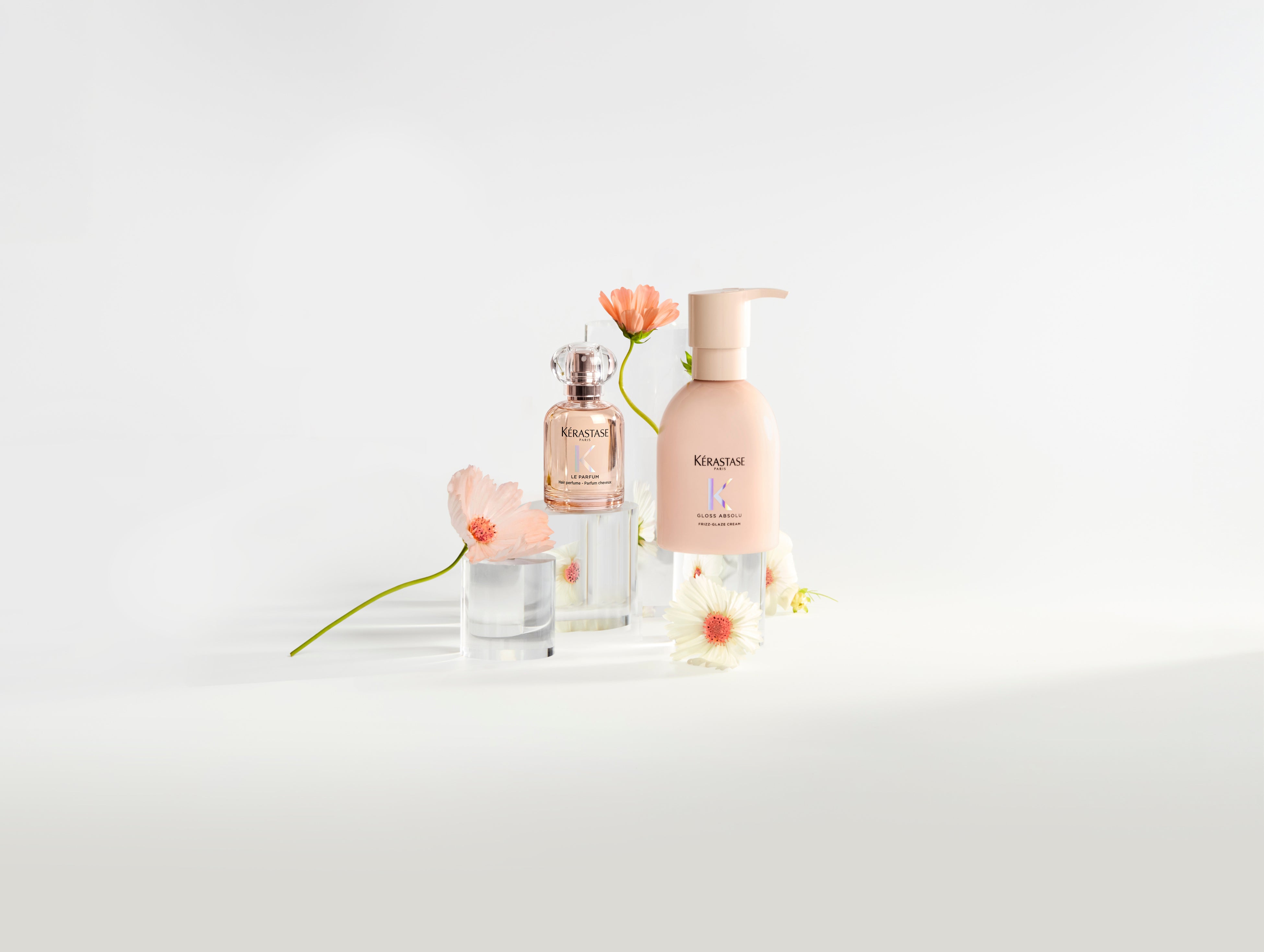 Kérastase Frizz-Glaze Cream and Le Parfum hair perfume on clear plinths with pale flowers.
