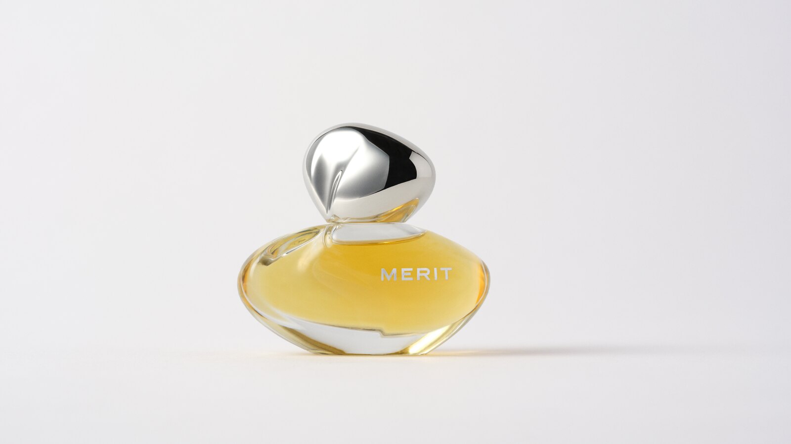 MERIT New Wave perfume in an asymmetric clear-glass bottle containing golden liquid, with a polished silver cap, on a pale grey background.